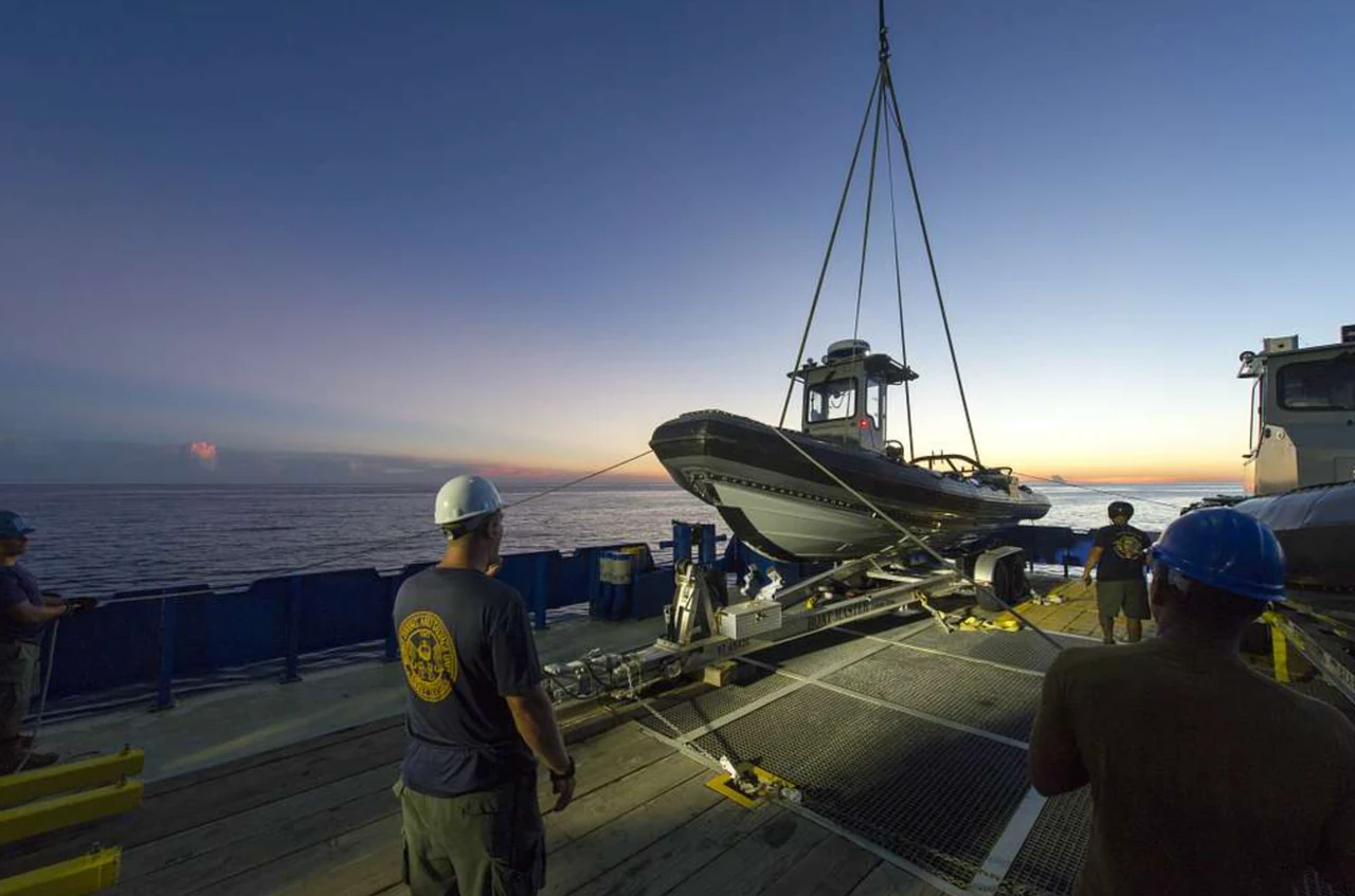 A rigid inflatable boat hangs from lifting slings above a ship’s deck at dusk, with crew members nearby.