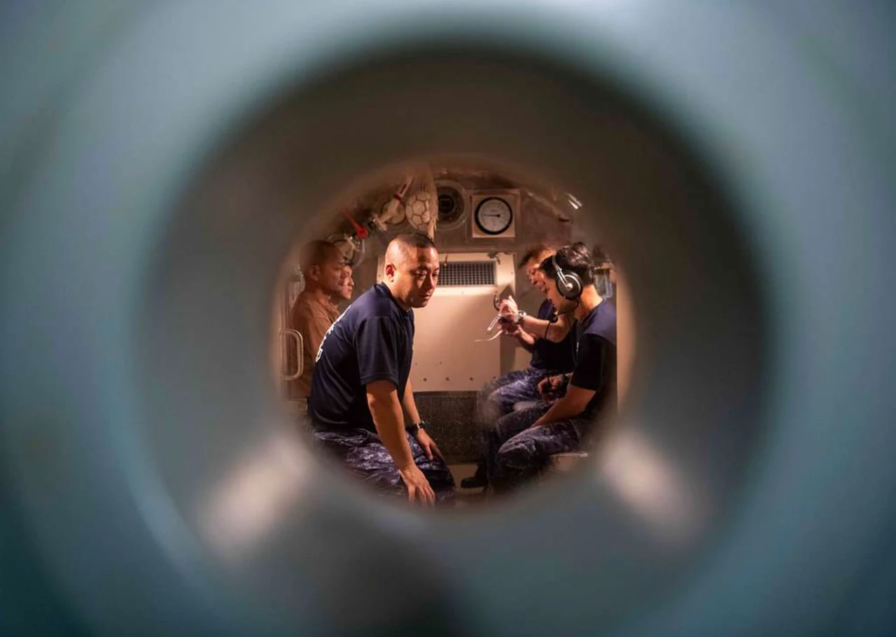 Four people seated inside a chamber, seen through a circular porthole.